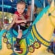 A smiling little boy with cerebral palsy rides on a colorful carousel horse as a smiling woman steadies him with her arms.